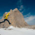 A hiker wearing a bright yellow jacket and a light green backpack jumps as they traverse a snowy mountain peak.