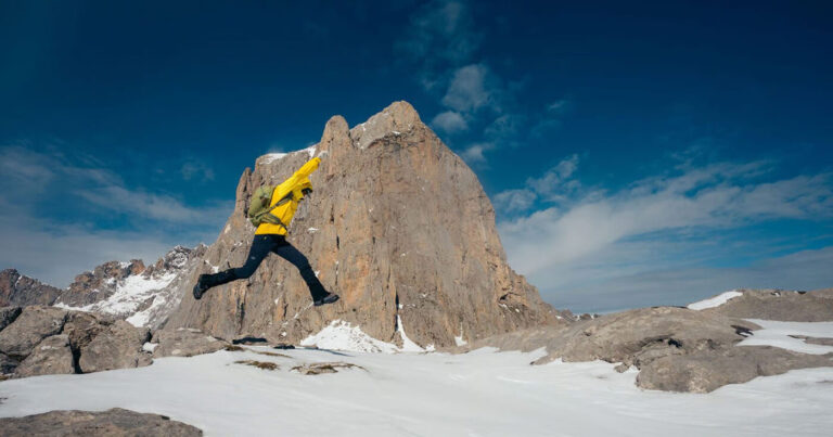 A hiker wearing a bright yellow jacket and a light green backpack jumps as they traverse a snowy mountain peak.
