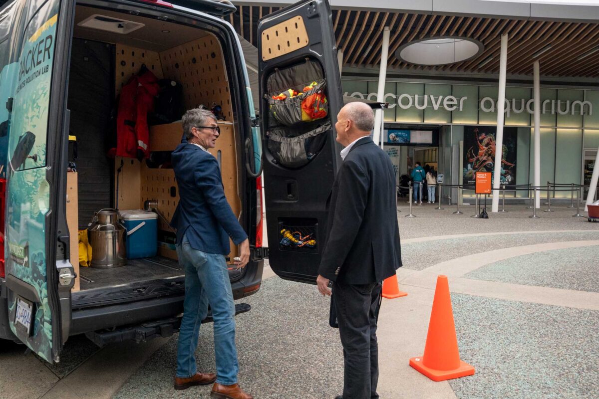 With the van parked outside the Vancouver Aquarium front entrance, the back doors of the van are open to reveal the renovated inside. Peter extends an arm to exhibit the design as Clint smiles and takes it in.
