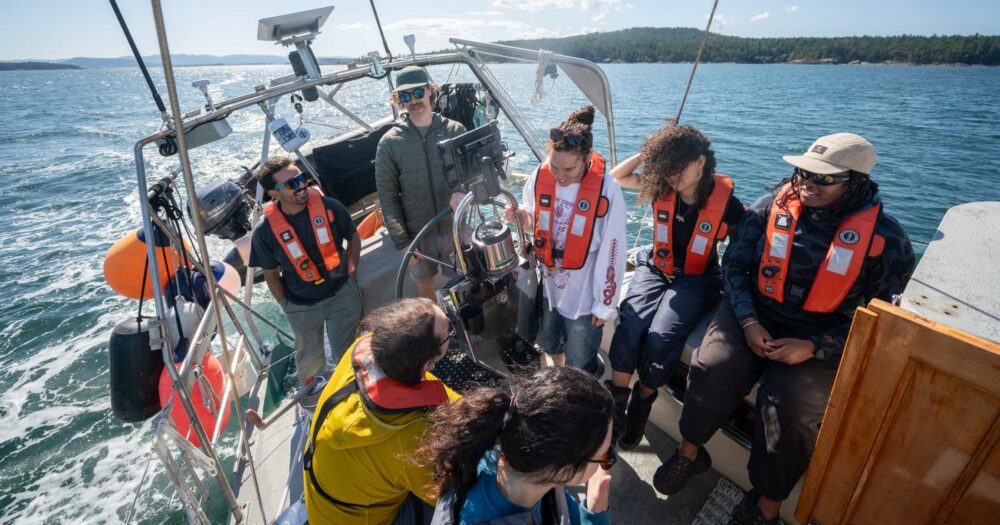 A group of 7 people sit/stand in a circle aboard Achiever.