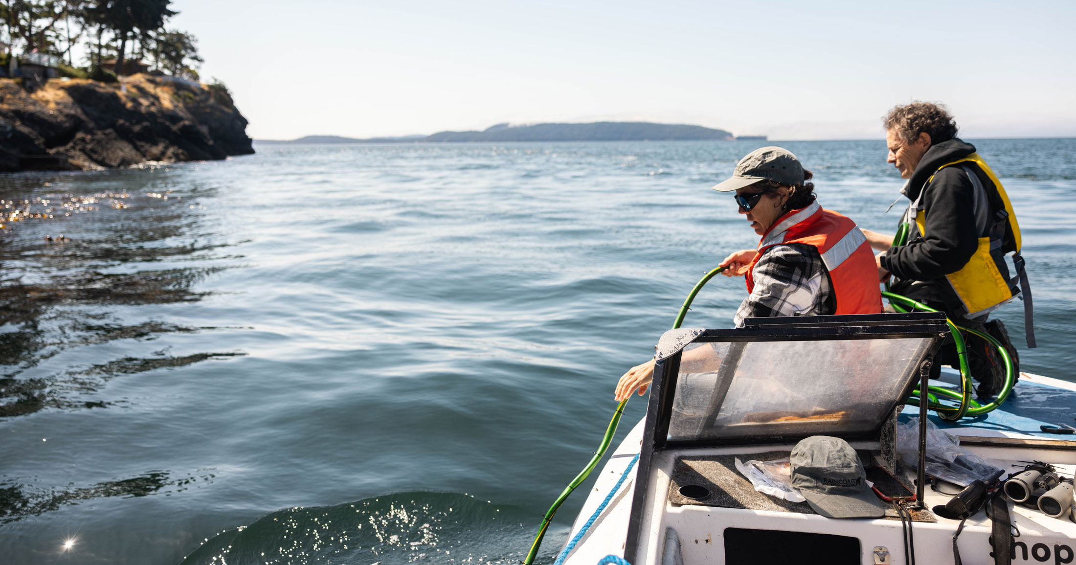 Two scientists appear on the right side of the image standing inside a small boat, placing a long wire into the ocean.