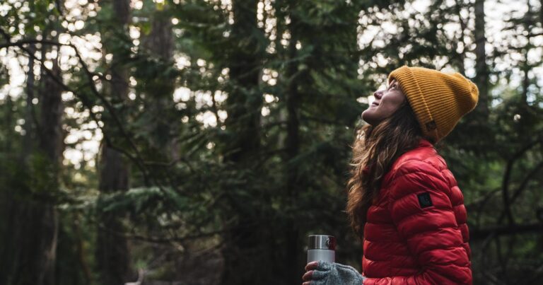 Shauna Doll stands with her winter coat and toque looking up in to the forest canopy, holding her hot drink.