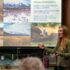 A red-haired young woman stands in front of a projector, speaking to a crowd sat listening as she speaks on wolf biology.