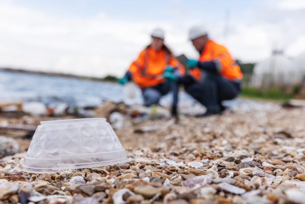 A plastic container lies on a polluted shoreline, with two environmental engineers in safety gear examining debris in the background. The image underscores pollution impact and the need for environmental action.