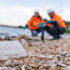 A plastic container lies on a polluted shoreline, with two environmental engineers in safety gear examining debris in the background. The image underscores pollution impact and the need for environmental action.