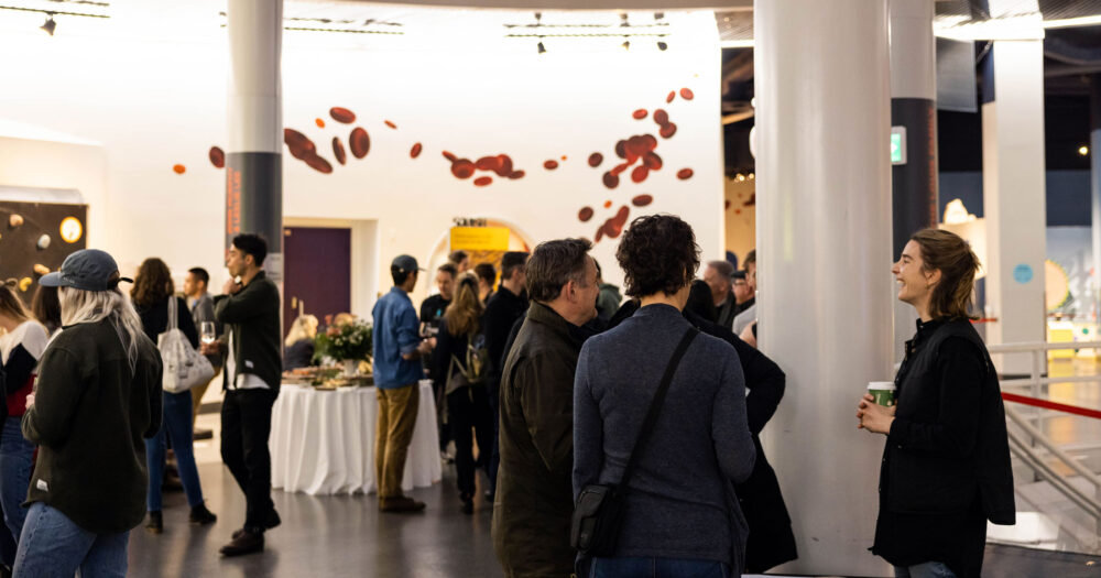 A group of young professionals gather in a large hall conversing with one another, laughing and enjoying beverages.