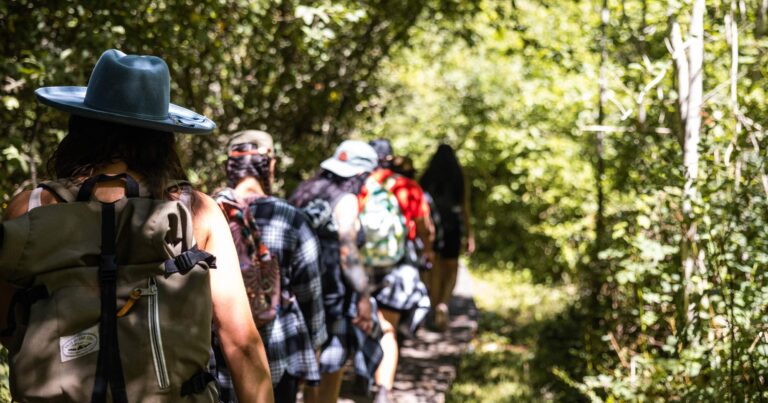 6 people walk in a single file line through the forest with their backs to the camera.