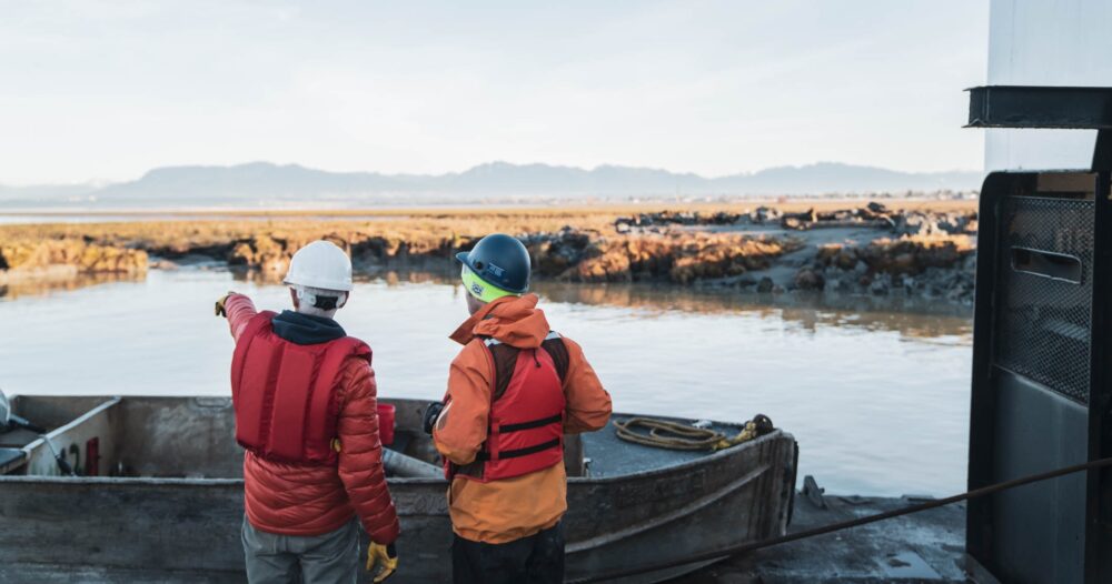 Two people wearing hard hats and life jackets face away from the camera and look out over the water.