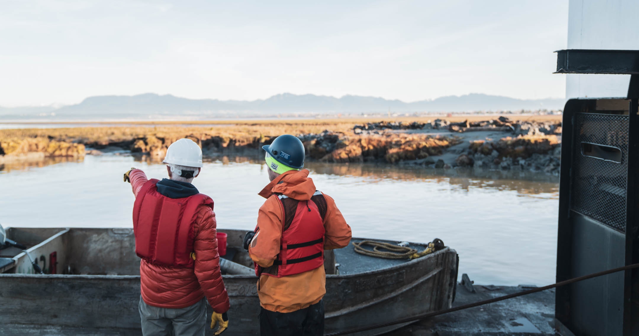 Two people wearing hard hats and life jackets face away from the camera and look out over the water.