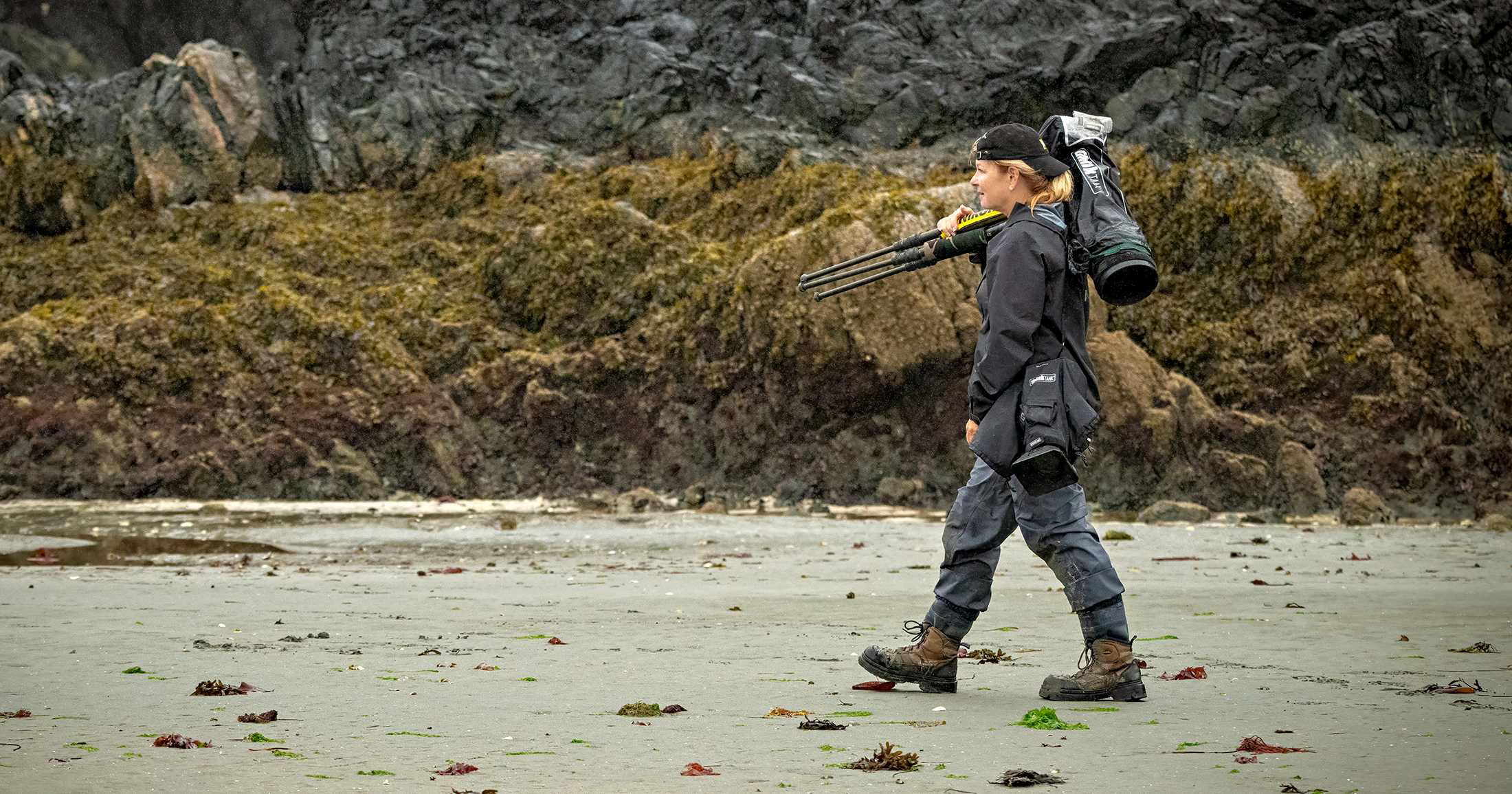 Michelle walks along a sandy beach wearing all black with a large camera and tripod over her shoulder.