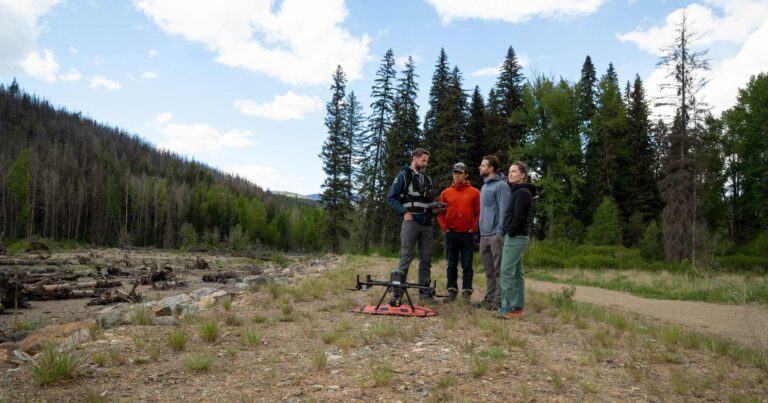 4 people stand outside around a drone near a riverbed.