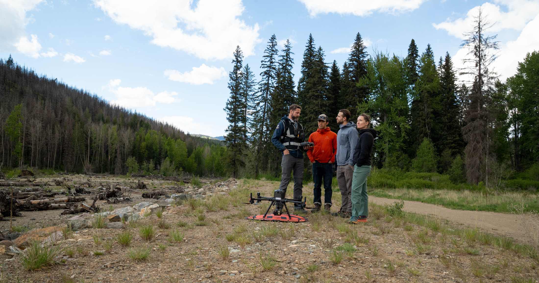 4 people stand outside around a drone near a riverbed.