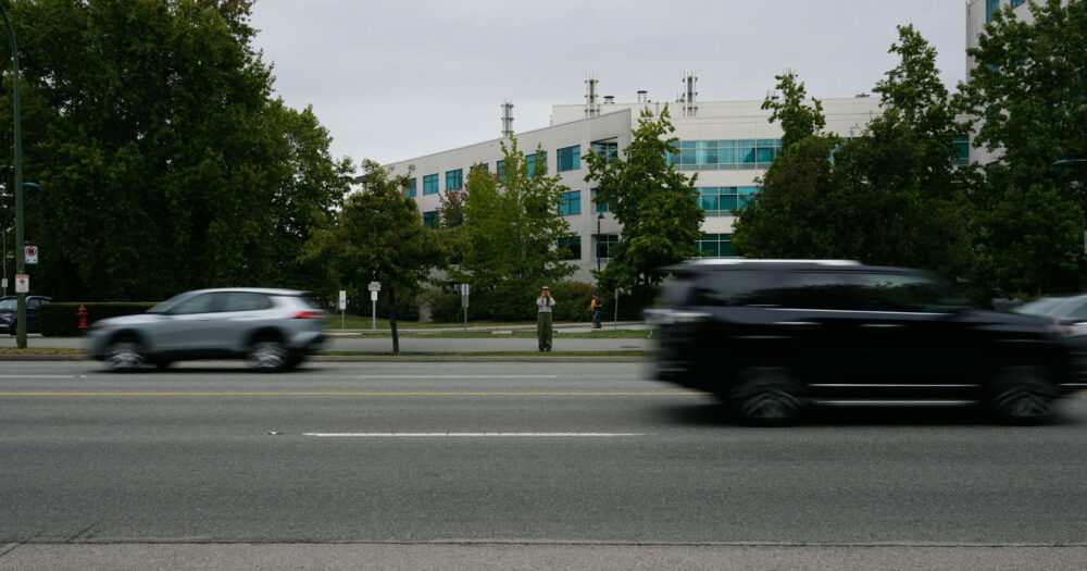 A woman stands across a busy street as cars zoom past, seemingly trying to yell something at the camera on the other side.