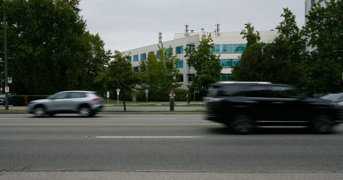 A woman stands across a busy street as cars zoom past, seemingly trying to yell something at the camera on the other side.
