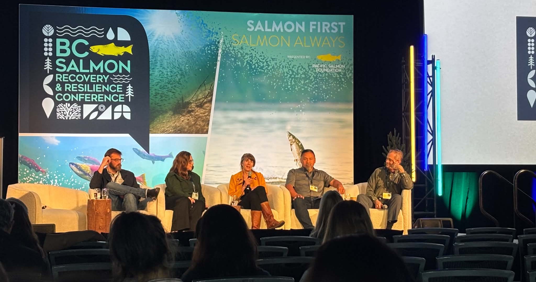 A panel of 5 people sit in chairs on stage, including Raincoast's Misty MacDuffee sitting in the middle.